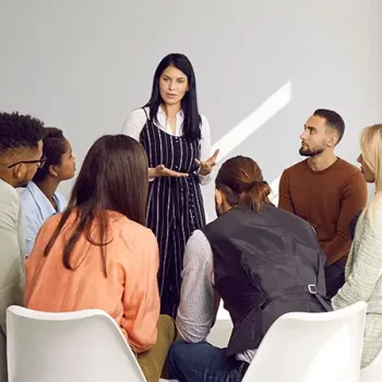 Small group of people sitting in a circle with a woman standing, addressing the group.